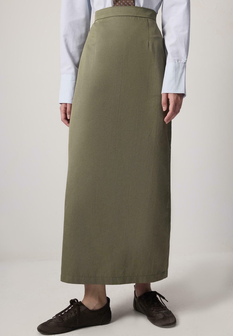 Olijfgroene midi rok van gladde stof met hoge taille en rechte snit, gecombineerd met een lichtblauw shirt en donkerbruine schoenen.