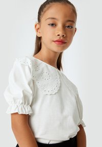Camisa blanca con un cuello con encaje, mangas cortas abullonadas y puños fruncidos. Textura suave, diseño simple y sin gráficos visibles.