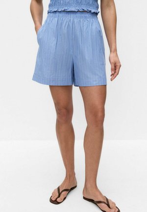 Kvinde iført lyseblå, stribede shorts med elastisk talje og sidelommer, kombineret med sorte flip-flops.