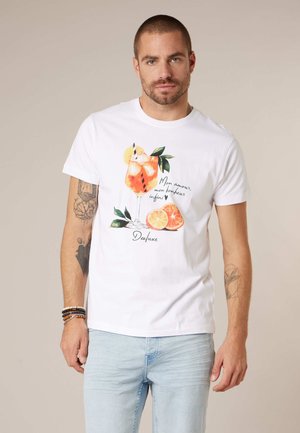 Man draagt een wit T-shirt met een afbeelding van een ijskoud sinaasdrankje, citrusvruchtenschijfjes en Franse tekst, gecombineerd met lichtblauwe jeans en kralenarmbanden.