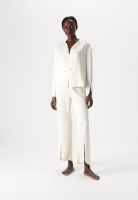Světlý krémový loungewear set vyrobený z texturované tkaniny, obsahující košilový top s límečkem a kalhoty po kotníky se bočními rozparky.