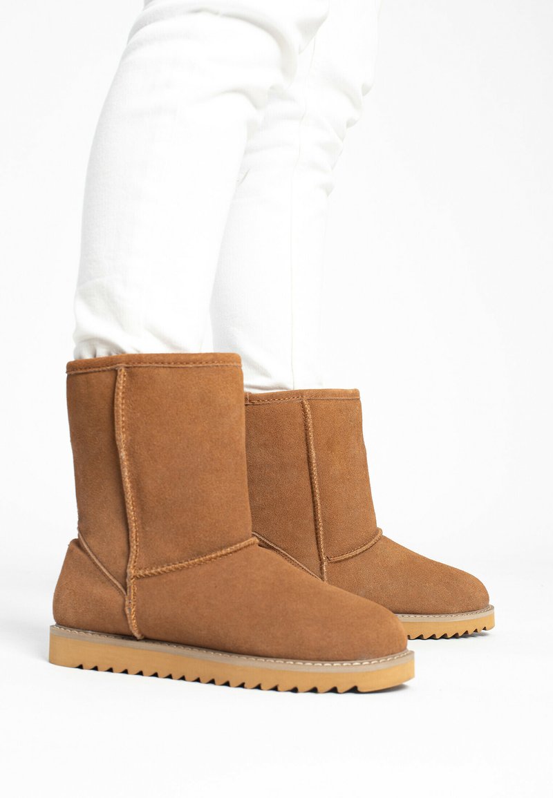 Botas de tobillo de ante color marrón claro con un tubo recto, suela plana y dibujo de zigzag. Las costuras son visibles, la textura parece suave y resistente.