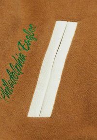 Il tessuto in feltro marrone presenta una zip in pelle bianca come dettaglio. Il testo ricamato in verde recita "Philadelphia Eagles" lungo il lato.