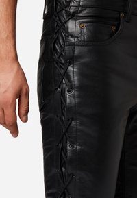 Zwarte leren broek met een vetersluiting aan de zijkant, voorzien van metalen oogjes, en voorzakken met goudkleurige accenten.