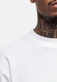 White waffle-knit sweatshirt with a round neckline, showing texture details and visible stitching. Tattoos are present on the neck area.