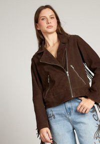 Brązowa skórzana kurtka krótkiej długości z wyciętym kołnierzem, zapinana na zamek z przodu, z dwoma bocznymi kieszeniami i frędzlami na rękawach. Noszona na jasnoniebieskich dżinsach.