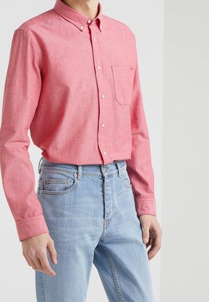 Homme portant une chemise rose à manches longues boutonnée, rentrée dans un jean en denim bleu clair sur un fond uni.