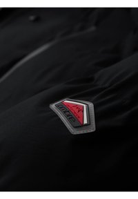 Primo piano di un tessuto nero con una toppa cucita che riporta le parole "performance" e "amture" nei colori rosso, bianco e nero.
