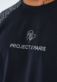 Camiseta negra con cuello redondo, mangas cortas y un patrón geométrico embozado en los hombros con "PROJECT X PARIS" en bordado plateado.
