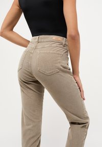 Beige corduroy high-waisted broek met een gestructureerde afwerking, twee achterzakken en een label op de tailleband. De pasvorm is op maat gemaakt en heeft een rechte pijp.