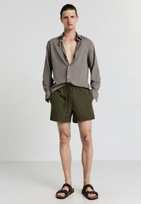 Graues Hemd mit Knopfleiste und offener Kragen, kombiniert mit olivgrünen Shorts mit Kordelzug und braunen Sandalen. Einfaches Design und Materialien.