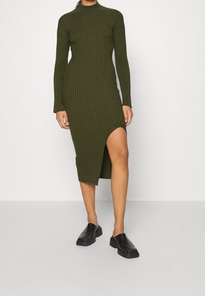Femme portant une robe midi en maille côtelée vert olive, à manches longues et fente haute sur le côté, associée à des chaussures noires sans lacets.