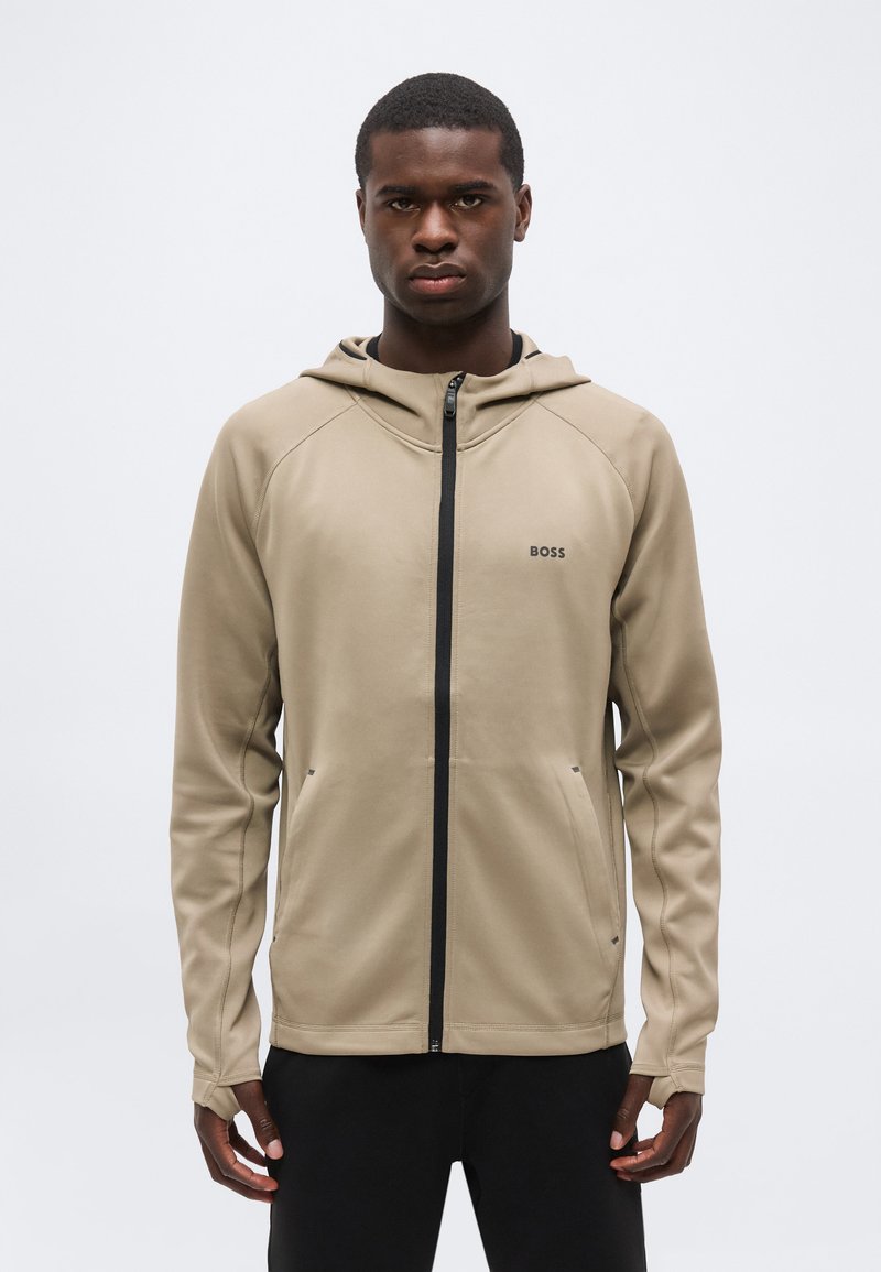 Jeune homme à l'expression sérieuse portant un sweat à capuche beige zippé avec une fermeture éclair noire et le logo "BOSS" sur la poitrine, debout devant un fond uni.