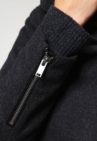 Sudadera negra con tela texturizada, primer plano del bolsillo con cremallera con herrajes plateados y puños acanalados para un ajuste ceñido.