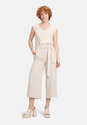 Beige jumpsuit med V-udskæring, ærmeløs design, selvbinder i taljen, brede ben, parret med glossy blokhælede sandaler.