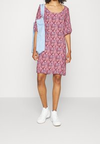 Červené a růžové vzorované šaty s krátkými balónovými rukávy, výstřihem do oblouku a délkou po kolena, spárované s světle modrou kabelkou přes rameno a bílými teniskami.