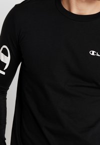 Schwarzes langärmeliges T-Shirt aus Baumwolle; mit einem weißen Logo auf der Brust und einer großen weißen Zahl am linken Ärmel.
