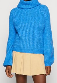 Pull à col roulé en maille épaisse bleu associé à une mini-jupe plissée beige porté par une personne à la peau foncée.