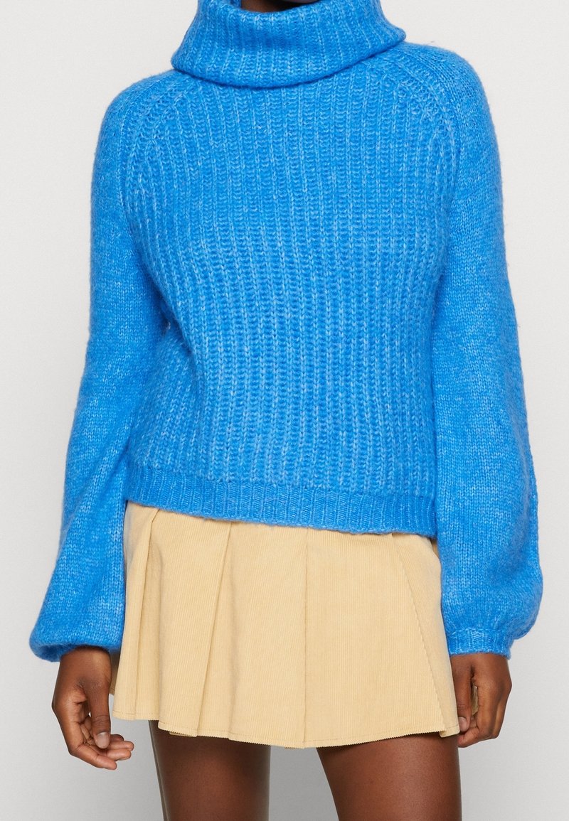 Pull à col roulé en maille épaisse bleu associé à une mini-jupe plissée beige porté par une personne à la peau foncée.