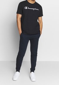 Muž v černém tričku Champion, tmavě modrých teplákách a bílých teniskách, stojící s rukama v kapsách na jednobarevném pozadí.