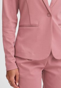 Svetle růžový přizpůsobený blazer s jedním knoflíkem, úzký střih a přední kapsy. Hladká tkanina s jemnou texturou.