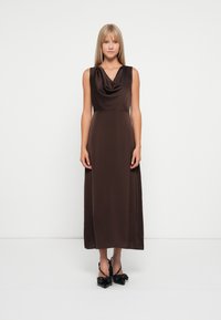 Robe sans manches en satin marron avec un col drapé en cascade et une silhouette longue et fluide. Portée avec des talons noirs à bout pointu.