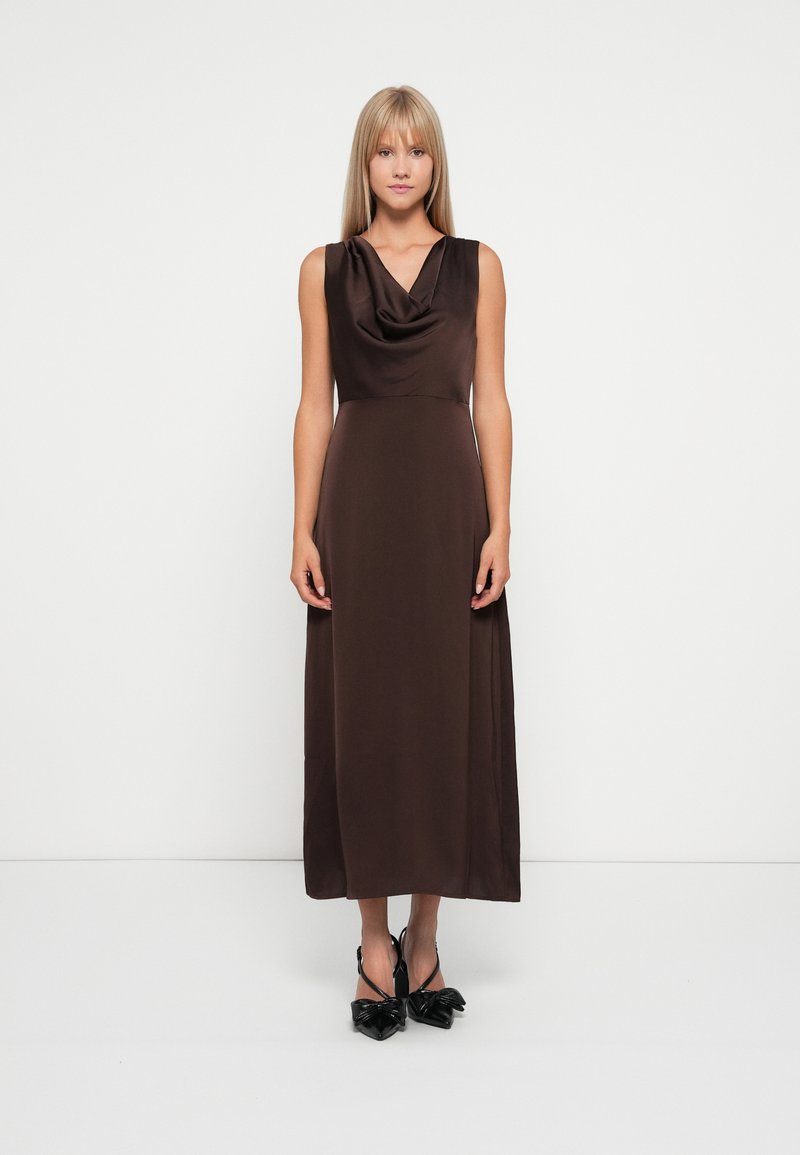 Robe sans manches en satin marron avec un col drapé en cascade et une silhouette longue et fluide. Portée avec des talons noirs à bout pointu.