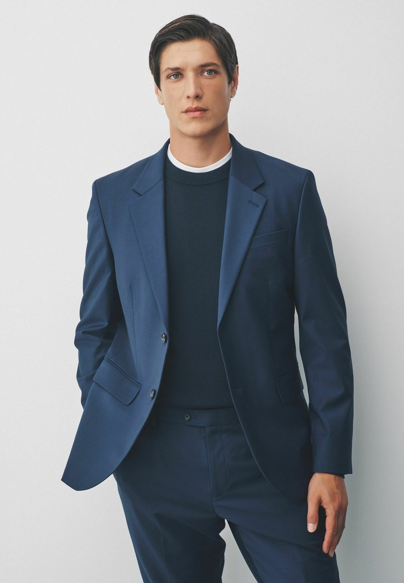 Marineblauw pak met een slim-fit, bestaande uit een enkelvoudig colbert en bijpassende broek. Daaronder een donkere trui met een witte kraag.