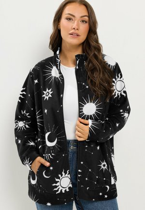 Femme portant une veste noire zippée avec des motifs blancs de soleil, lune et étoiles, associée à un top blanc et un jean bleu.