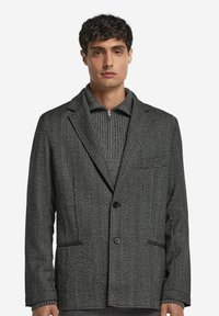 Grauer Fischgrat-Blazer mit Revers, zwei vorderen Taschen und einem einzelnen Knopfverschluss, kombiniert mit einem grauen gerippten Pullover.