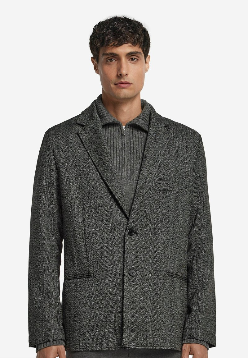 Grauer Fischgrat-Blazer mit Revers, zwei vorderen Taschen und einem einzelnen Knopfverschluss, kombiniert mit einem grauen gerippten Pullover.