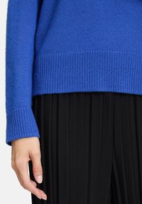 Main gauche à côté d'une jupe plissée noire, portant un pull en tricot bleu vif avec des poignets et un ourlet côtelés.