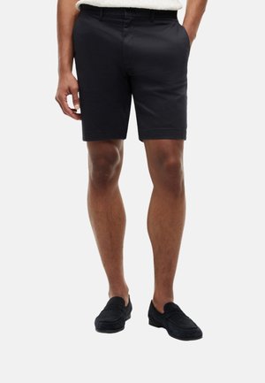 Hombre vestido con pantalones cortos negros a medida y zapatos negros tipo slip-on, de pie con las manos parcialmente en los bolsillos frente a un fondo blanco.