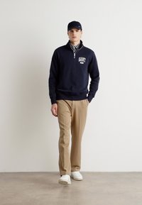 Hanorac cu fermoar bleumarin cu logo, pantaloni bej și adidași albi. Modelul poartă o șapcă bleumarin asortată și o cămașă în carouri dedesubt.