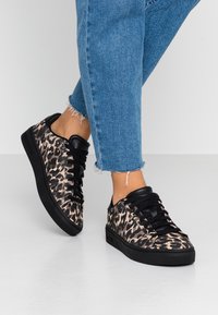 Tenisky s leopardím vzorem mají černý texturovaný svršek, šněrování a černou gumovou podrážku, a jsou kombinovány s modrými džíny s roztrhaným lemem.