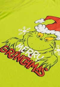 T-shirt verde brillante con un Grinch in cartoon che indossa un cappello da Babbo Natale, con il testo "MERRY GRINCIMAS" in rosso e bianco. Tessuto di cotone morbido.
