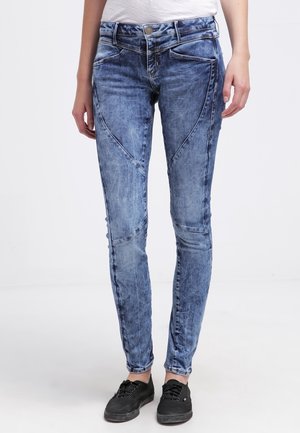 Slim fit blå acid-wash jeans med dekorative syninger båret med sorte sneakers på en stående person mod en ensfarvet baggrund.