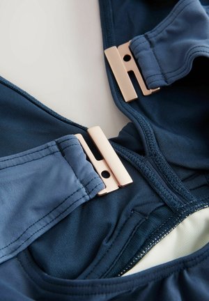 Costume da bagno blu navy realizzato in tessuto liscio e elastico. Presenta chiusure in metallo rosa dorato e una texture interna a contrasto.