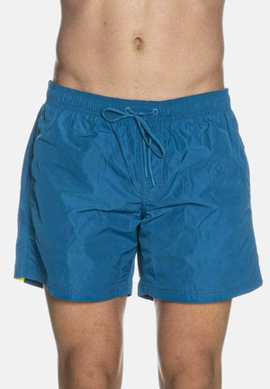 Uomo che indossa pantaloncini da bagno blu con elastico in vita e coulisse, mostrando l'addome nudo e la parte superiore delle cosce su uno sfondo uniforme.