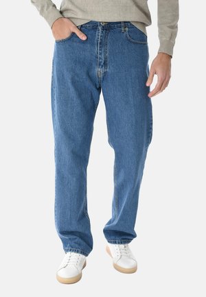 Uomo con jeans blu a gamba dritta, maglione beige e scarpe da ginnastica bianche, con una mano in tasca, su sfondo bianco.