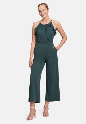 Tuta verde scuro con spalline arricciate, design a gamba larga e tasche laterali. Il tessuto morbido cade in modo elegante, abbinato a tacchi beige chiaro.