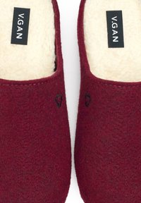Burgundy felt slip-on slippers with a soft cream lining, featuring a black logo tag and subtle dark accents on the outer surface.
