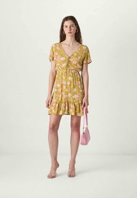 Vestido floral amarelo com mangas curtas, decote em V profundo, cintura com laço e barra rodada. A modelo segura uma pequena bolsa rosa e usa sandálias transparentes.