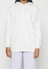 Sudadera blanca con capucha ajustable, puños acanalados y un logo en el pecho. Textura suave, ajuste relajado, combinada con pantalones de pierna ancha en color lavanda.