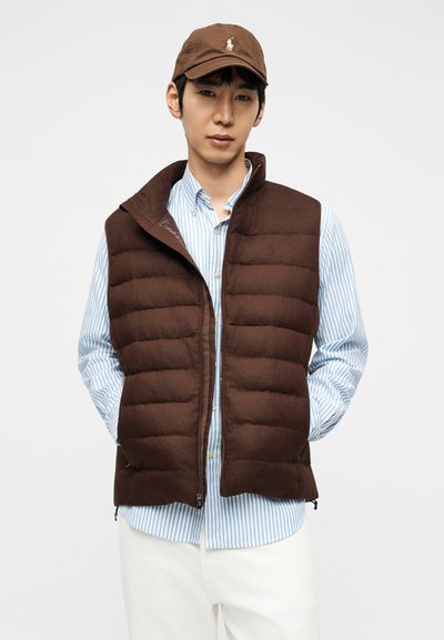 Νέος άνδρας με καφέ επενδυμένο γιλέκο πάνω από μπλε πουκάμισο με ρίγες, λευκό παντελόνι και καφέ καπέλο μπέιζμπολ, στέκεται με τα χέρια στις τσέπες.