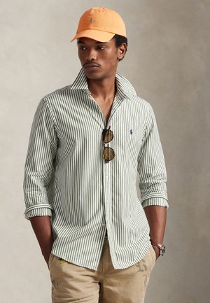 Camicia a righe verde e bianca con bottoni e colletto, indossata con pantaloni beige. Cappellino arancione e occhiali da sole come accessori. Design classico.