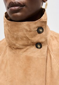 Κοντινό πλάνο ενός ατόμου που φοράει ένα μπεζ σατέν μπουφάν με ψηλό γιακά που ασφαλίζει με δύο μαύρα κουμπιά και ένα χρυσό σκουλαρίκι κρίκο.