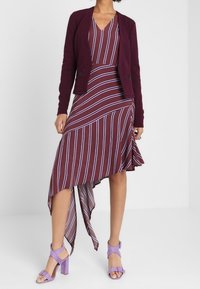 Abito bordeaux con righe bianche e lavanda, orlo asimmetrico, abbinato a un cardigan aderente coordinato e sandali con tacco viola.