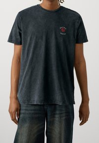 Camiseta negra de cuello redondo hecha de algodón lavado y suave, con un gráfico de una rosa roja y texto en el pecho. Corte estándar con mangas cortas.