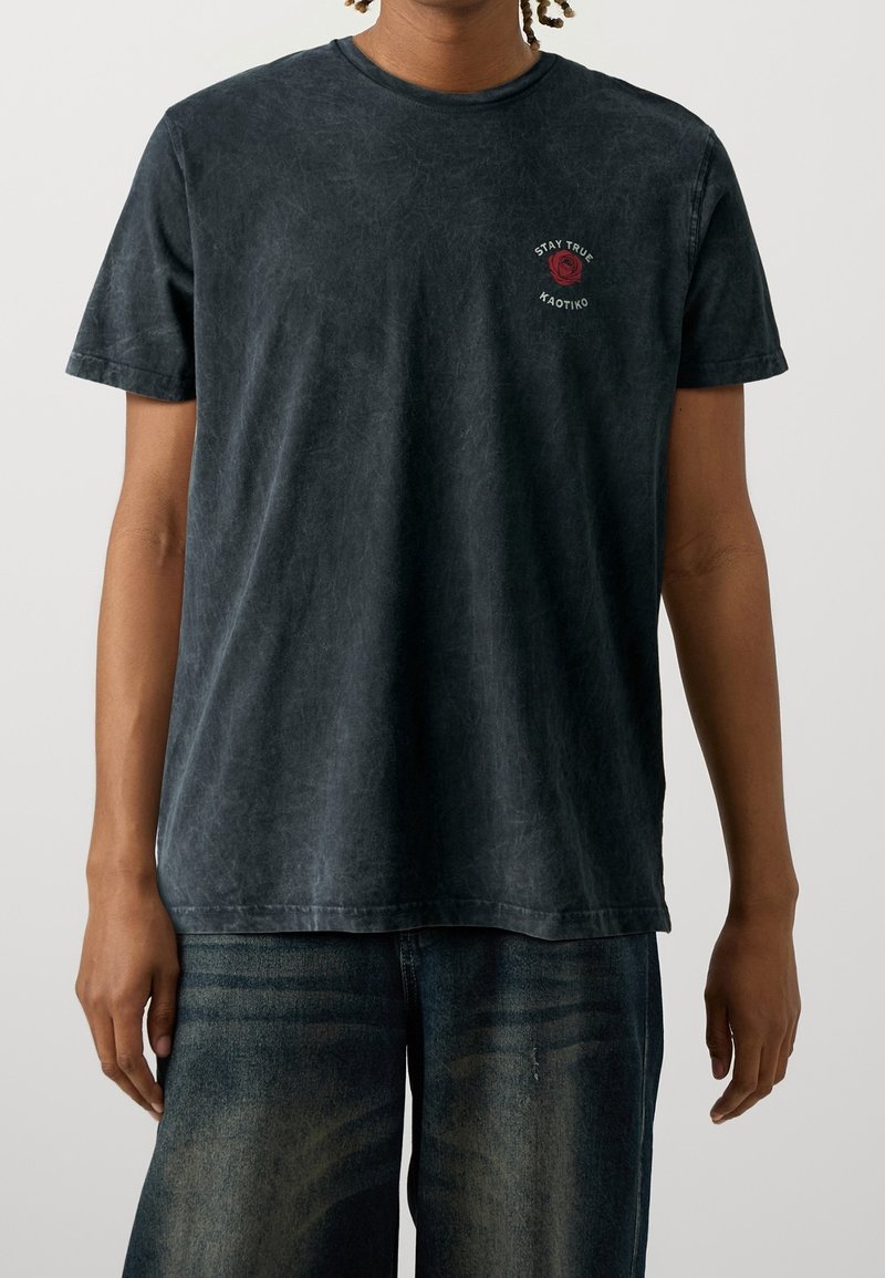 Camiseta negra de cuello redondo hecha de algodón lavado y suave, con un gráfico de una rosa roja y texto en el pecho. Corte estándar con mangas cortas.